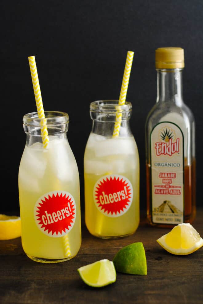Two small glass bottles with "cheers" labels on them, filled with a yellow liquid, ice, and decorative paper straws. A bottle of agave nectar is in background.