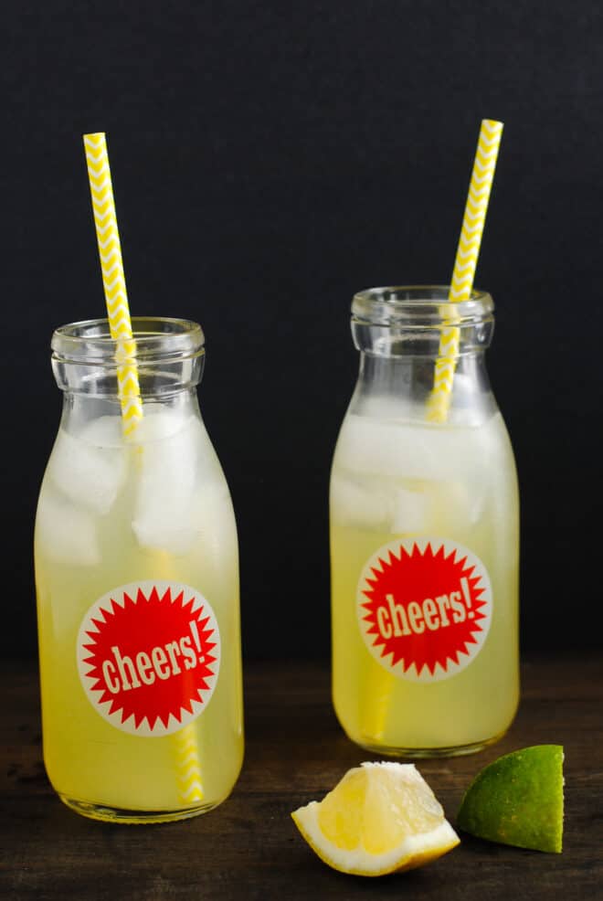 Two small glass bottles with red "cheers" stickers on them, filled with a yellow hued iced beverage and yellow straws.