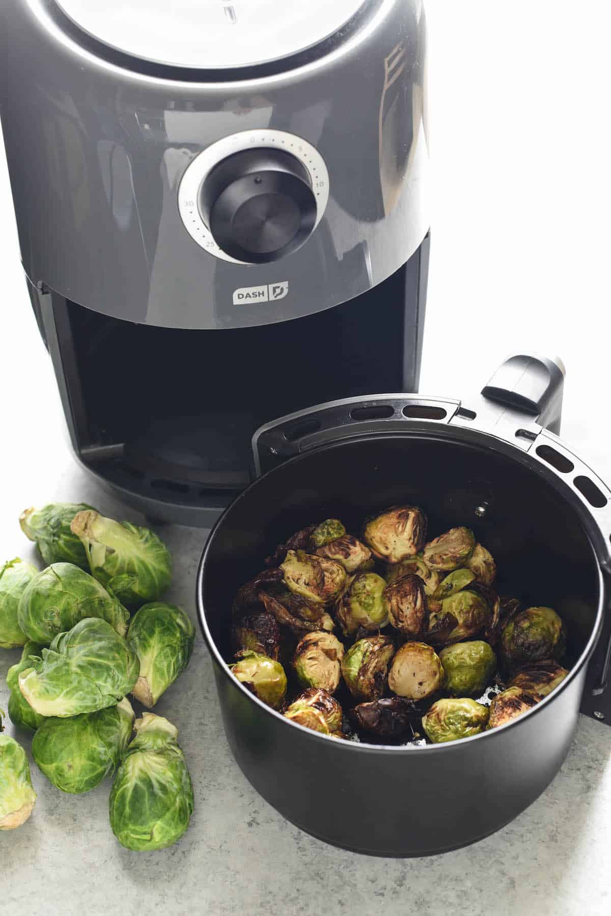 Brussel sprouts in air fryer basket with raw brussels sprouts on table.
