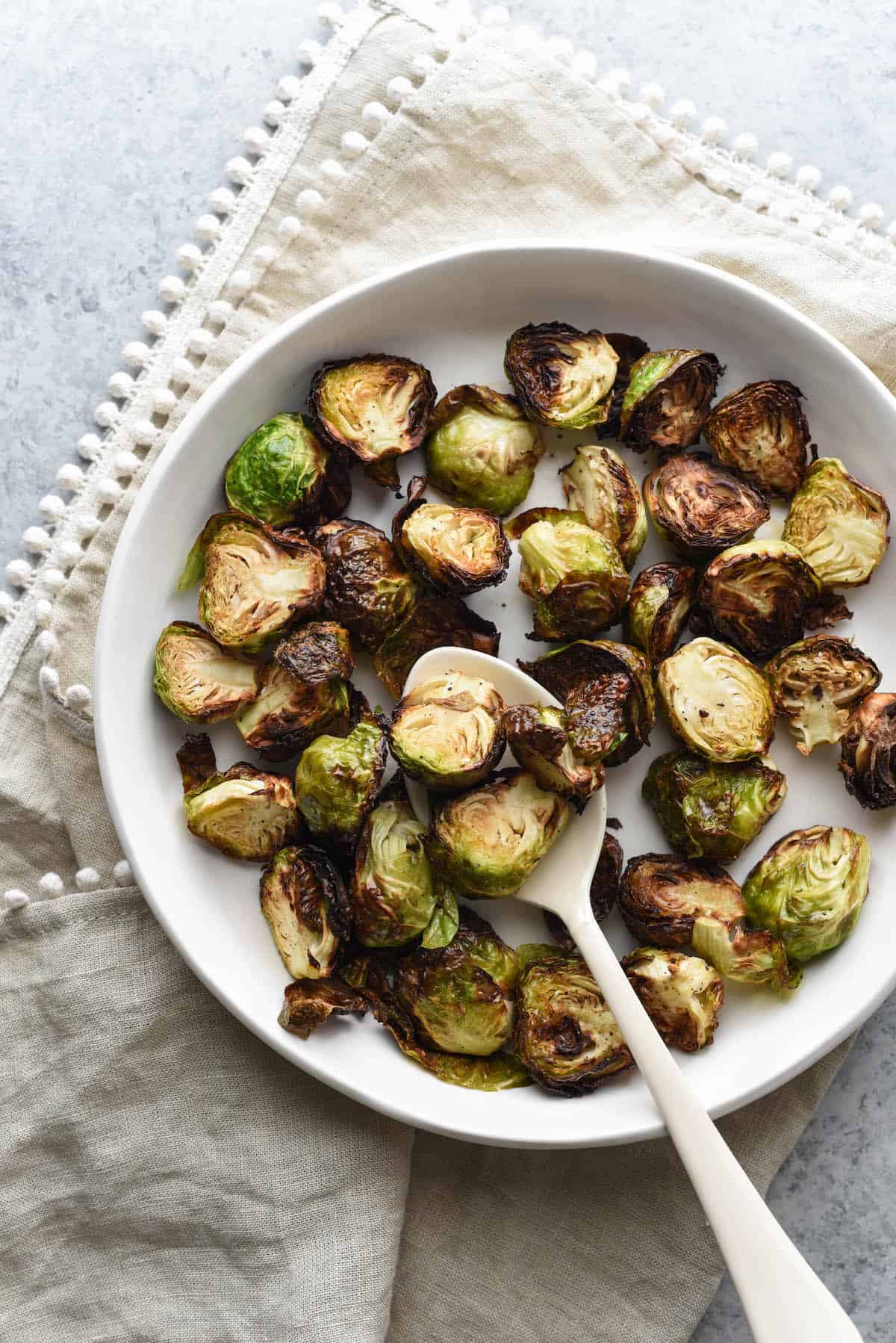 Air fry brussel sprouts in shallow white bowl with white serving spoon, on top of beige linen with pom pom edging.