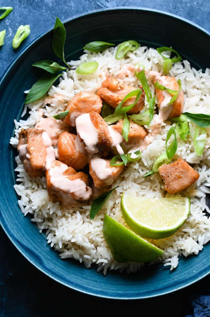 A blue bowl filled with white rice, air fryer salmon bites topped with a creamy sauce, sliced green onions and lime wedges.