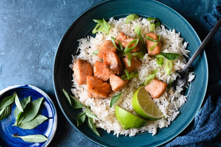 A blue bowl filled with white rice, air fryer salmon bites, sliced green onions and lime wedges, with a fork sticking into the rice.