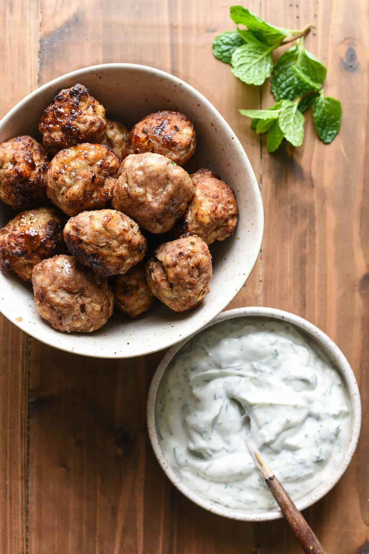A bowl of turkey meatballs air fryer and a bowl of herbed yogurt sauce, with a mint sprig.