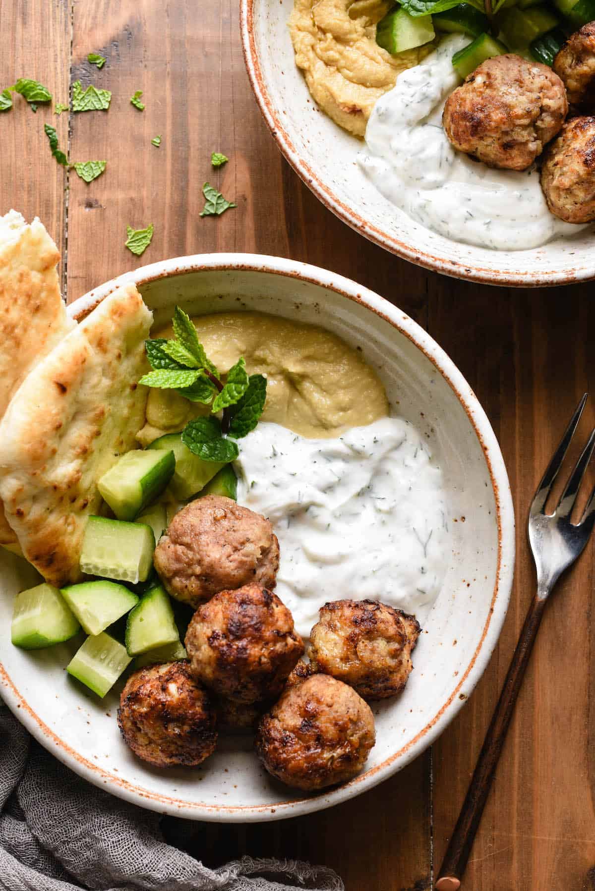 Two bowls filled with turkey meatballs in air fryer, diced cucumbers, herbed yogurt sauce, hummus and naan bread.