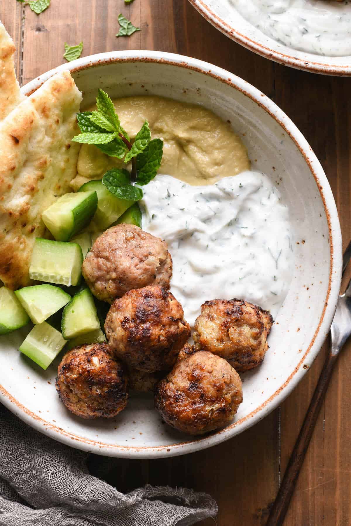 Overhead photo of rustic bowl filled with air fryer turkey meatballs, chopped cucumber, hummus, yogurt sauce, naan and a mint sprig garnish.