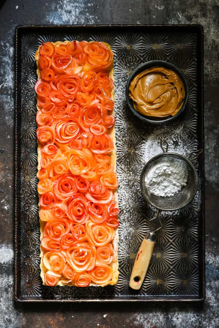 Rectangular tart topped with apple "roses" on vintage baking pan with bowl of peanut butter and powdered sugar sifter.