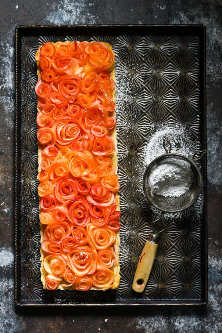 Long rectangle apple rose tart on vintage baking pan with powdered sugar sifter.
