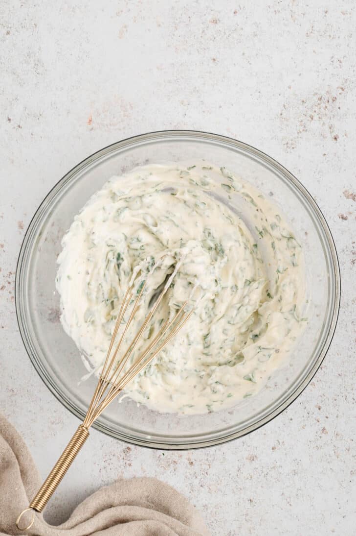 Cilantro mayonnaise being stirred with a whisk in a glass bowl on a light textured surface.