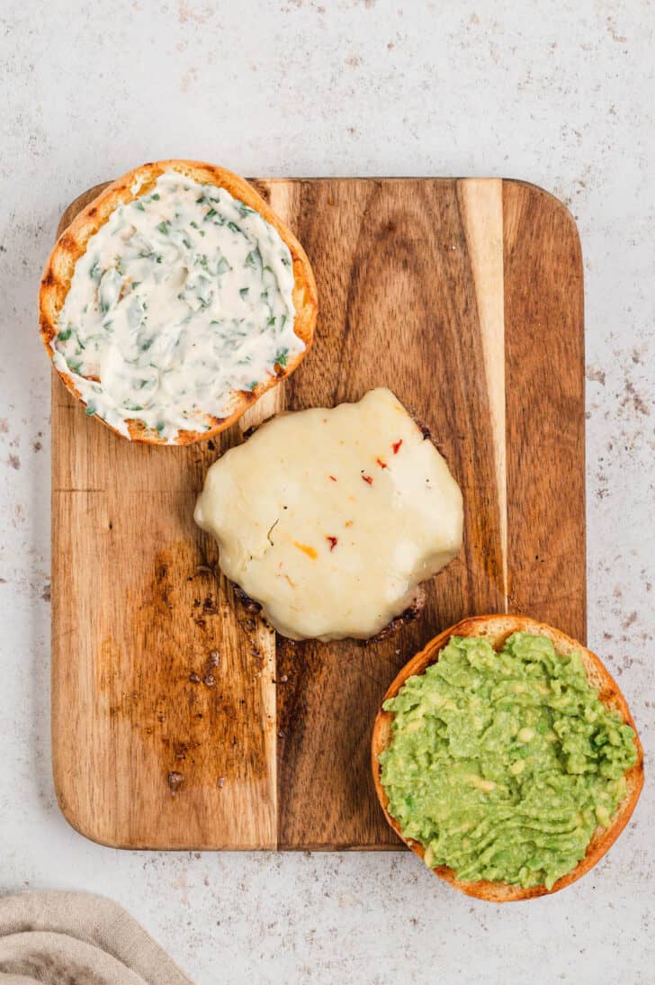 Two hamburger bun halves and a hamburger patty in a diagonal line on a wooden cutting board. One bun half is spread with cilantro mayo, and the other bun half has mashed avocado on it. The burger patty has melted pepper jack cheese.