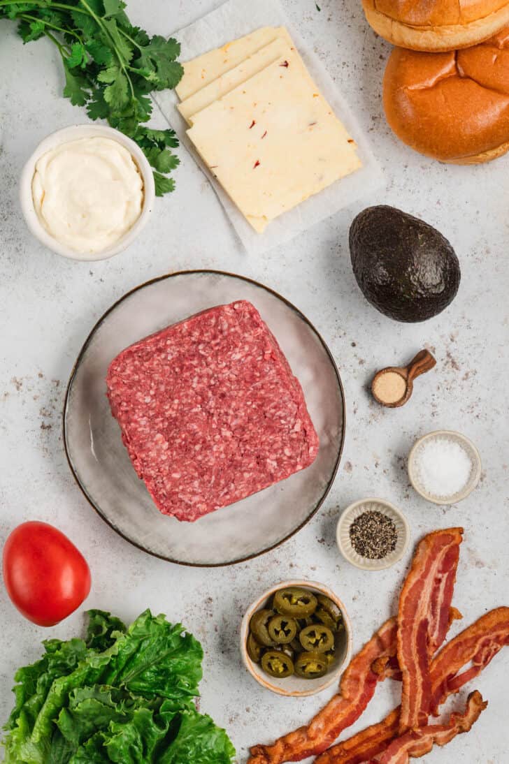 Ingredient laid out on a light textured surface, including a block of ground beef, an avocado, lettuce, tomato, cilantro, pickled jalapenos, bacon, mayonnaise, pepper jack cheese slices and spices.