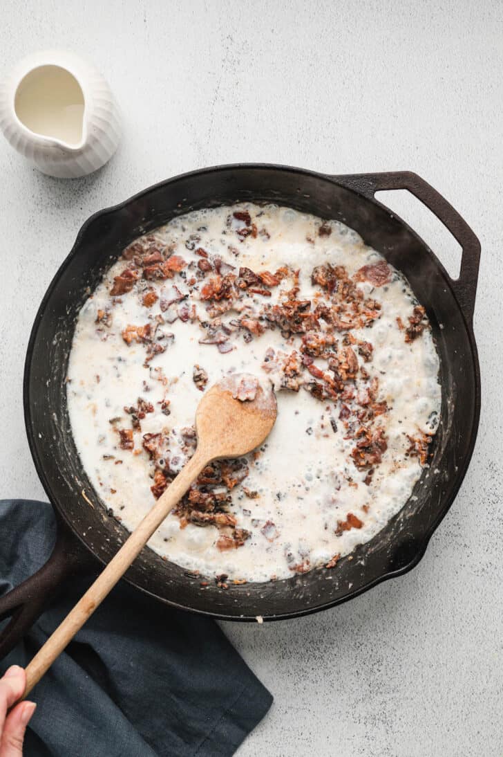 A mixture of milk and fried pork bits being stirred with a wooden spoon in a cast iron skillet.