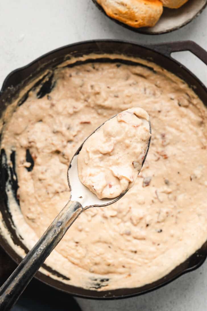 A spoon holding up a light brown creamy sauce over a cast iron skillet.