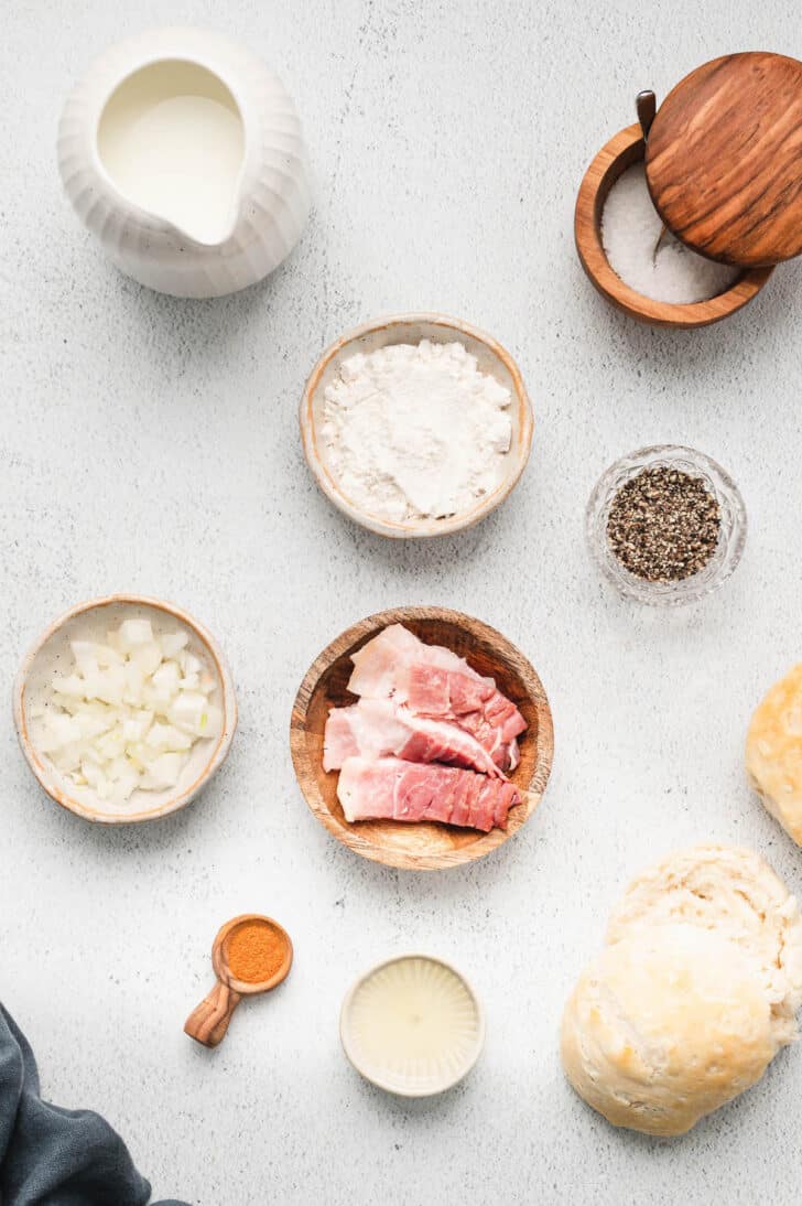 Ingredients laid out in small bowls on a white surface, including meat, onions, flour, biscuits and spices.