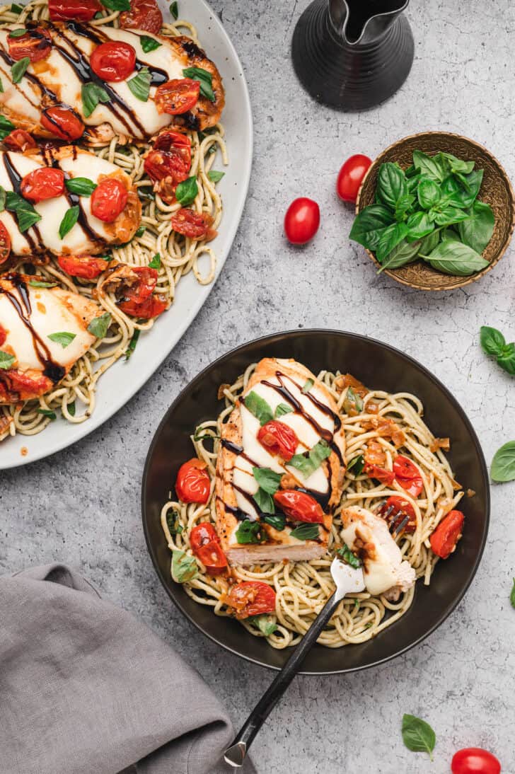 A black bowl filled with spaghetti topped with a chicken breast covered in mozzarella cheese, tomatoes, basil and balsamic glaze.