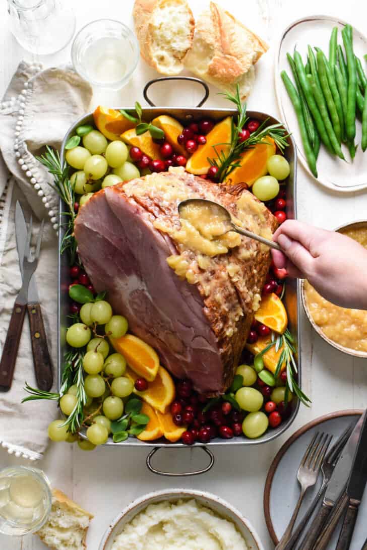 Metal roasting pan with spiral sliced ham surrounded by fruit garnishes, with woman's hand spooning pineapple sauce over ham.