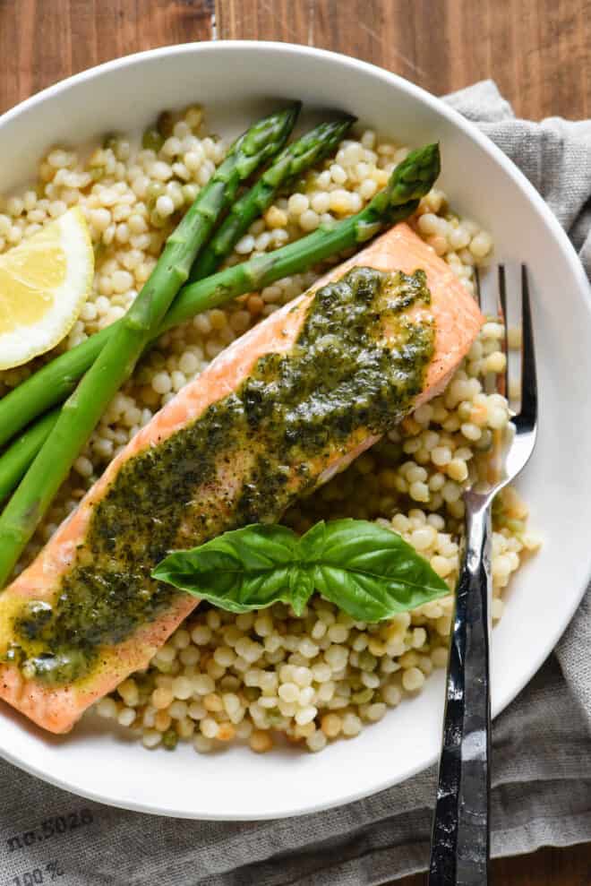 Shallow white bowl with pesto salmon, pearl couscous, asparagus spears and a lemon wedge.