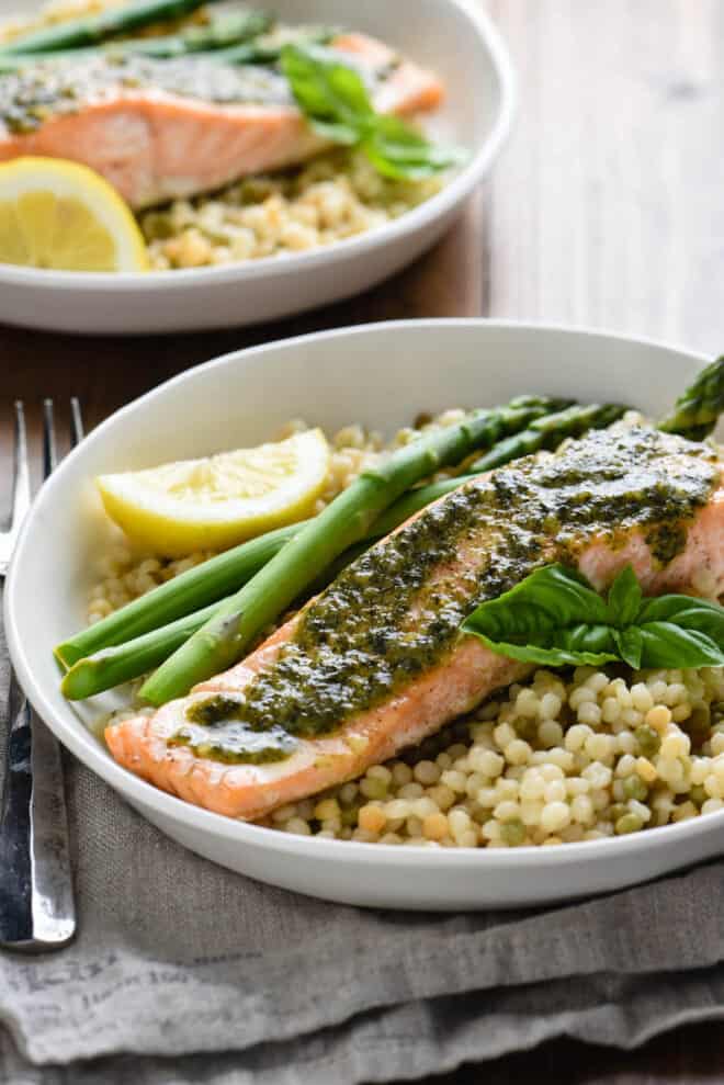 Shallow white bowl with baked fish topped with green herb sauce, tricolor couscous, asparagus, fresh basil sprig and lemon wedge.
