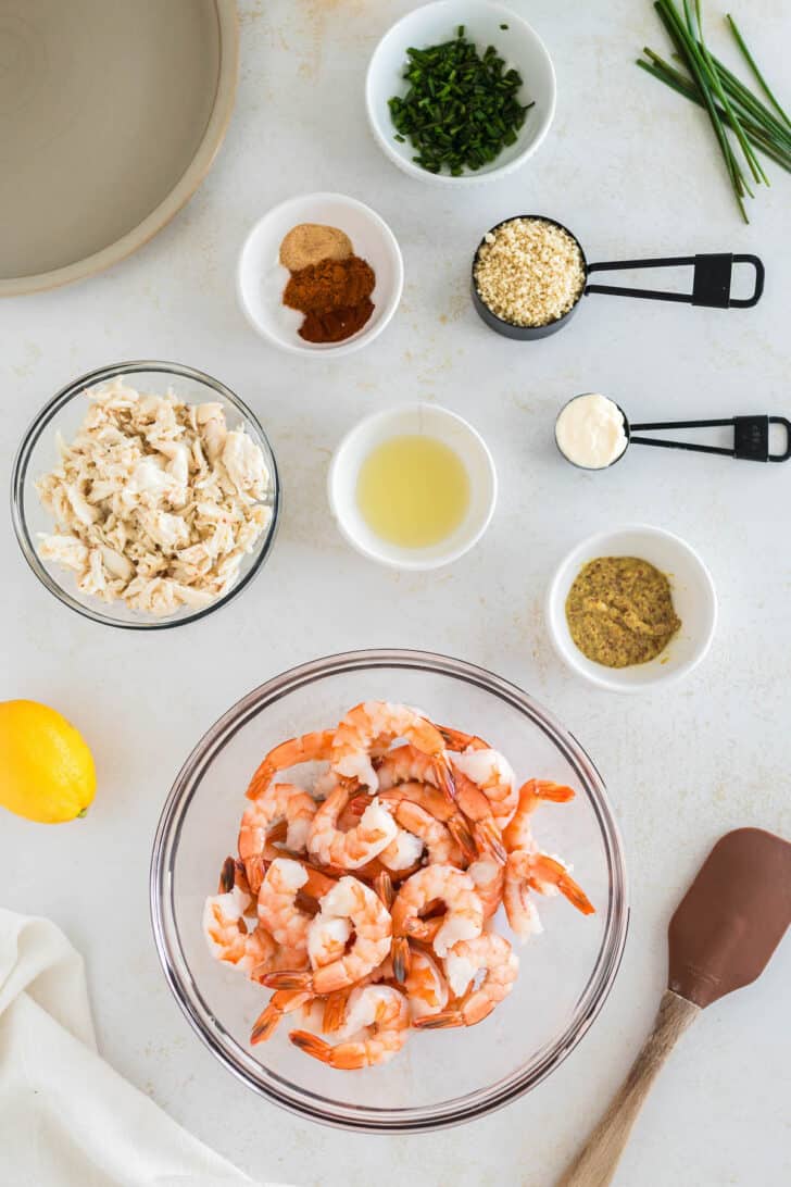 Ingredients laid out on a light surface, including spices, herbs, mayo, lemon juice and seafood.