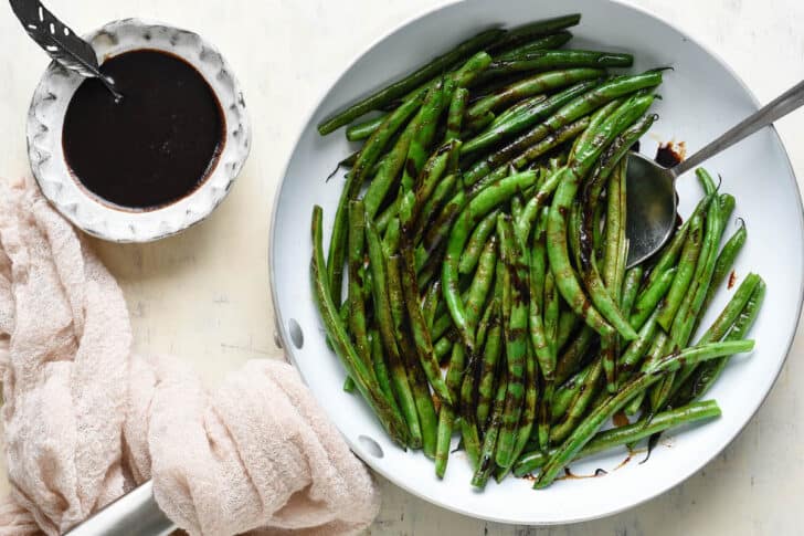 Balsamic glazed gren beans in a white skillet with a pink linen and bowl of glaze nearby.