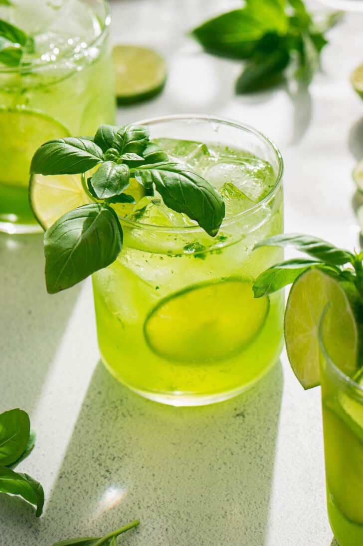 A green hued basil vodka cocktail garnished with a lemon wheel and fresh basil sprig.