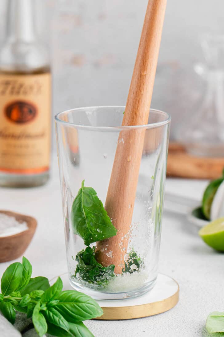 A wooden muddler crushing fresh herbs and sugar in a tall glass.