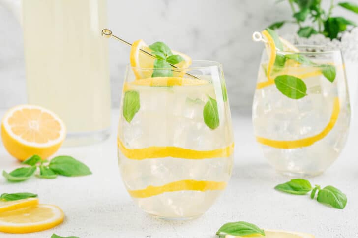 Two glasses filled with a basil lemonade cocktail, garnished with lemon peels and fresh basil leaves.