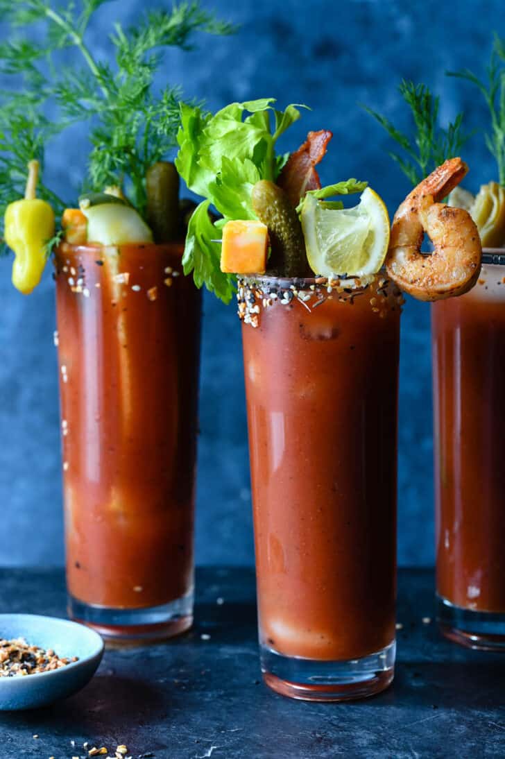 Three tall glasses of tomato juice with bloody mary garnish skewers including shrimp, lemon wedges, cheese and pickles.