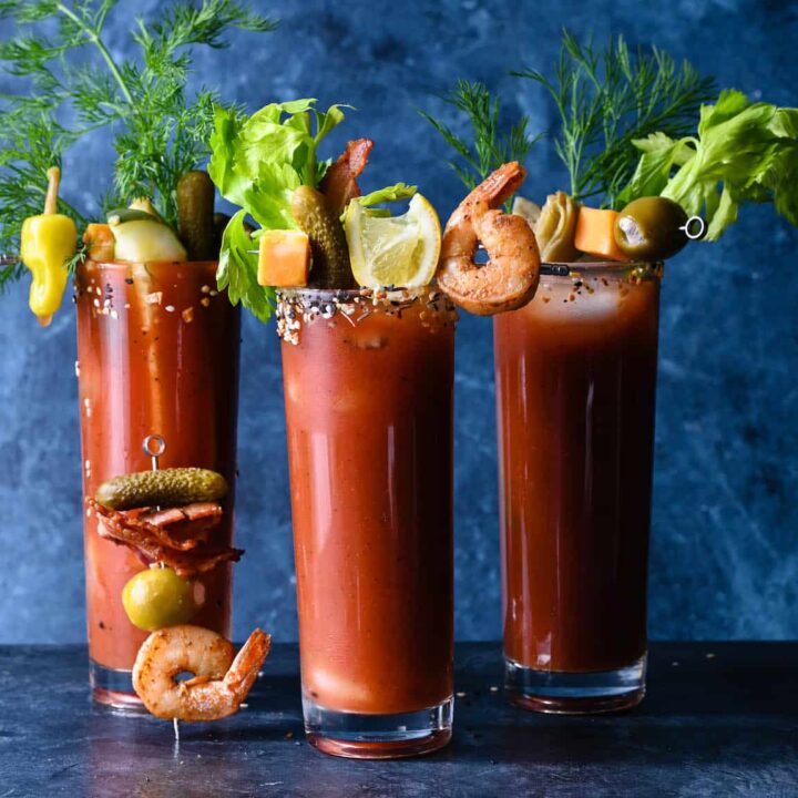 Three tall glasses filled with red juice, garnished with Bloody Mary skewers made with shrimp, bacon, cheese, pickles, artichokes and olives.