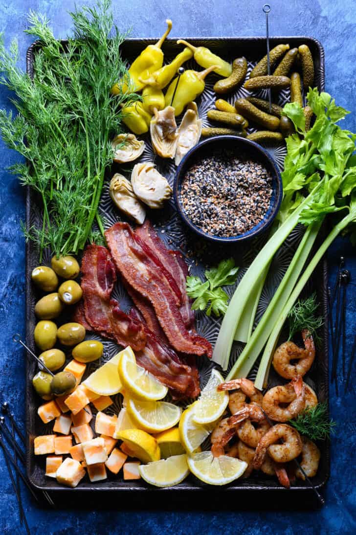 A textured baking pan topped with cheese cubes, green olives, bacon slices, fresh dill, celery, roasted shrimp, a bowl of everything bagel seasoning, artichoke hearts, pepperoncini and lemon wedges.