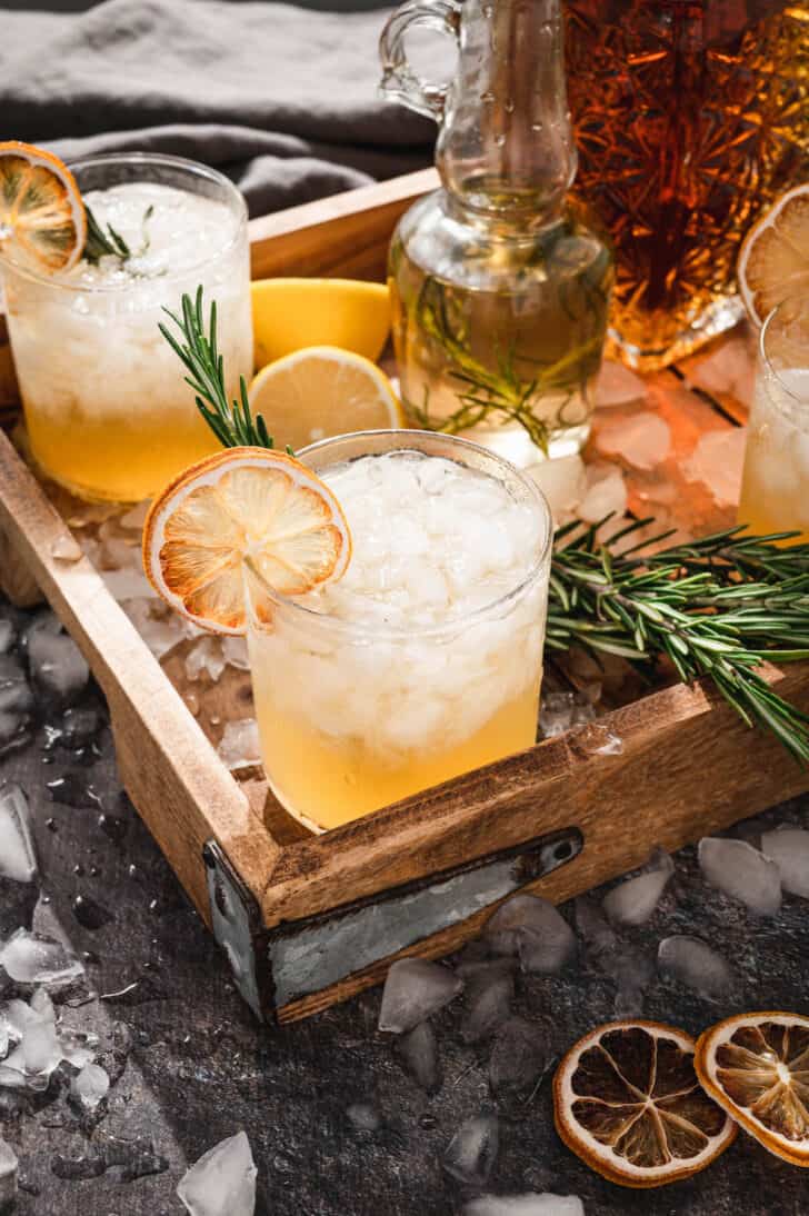 Lemon bourbon cocktails garnished with rosemary and dried citrus on a wooden tray with ice cubes all around the scene.