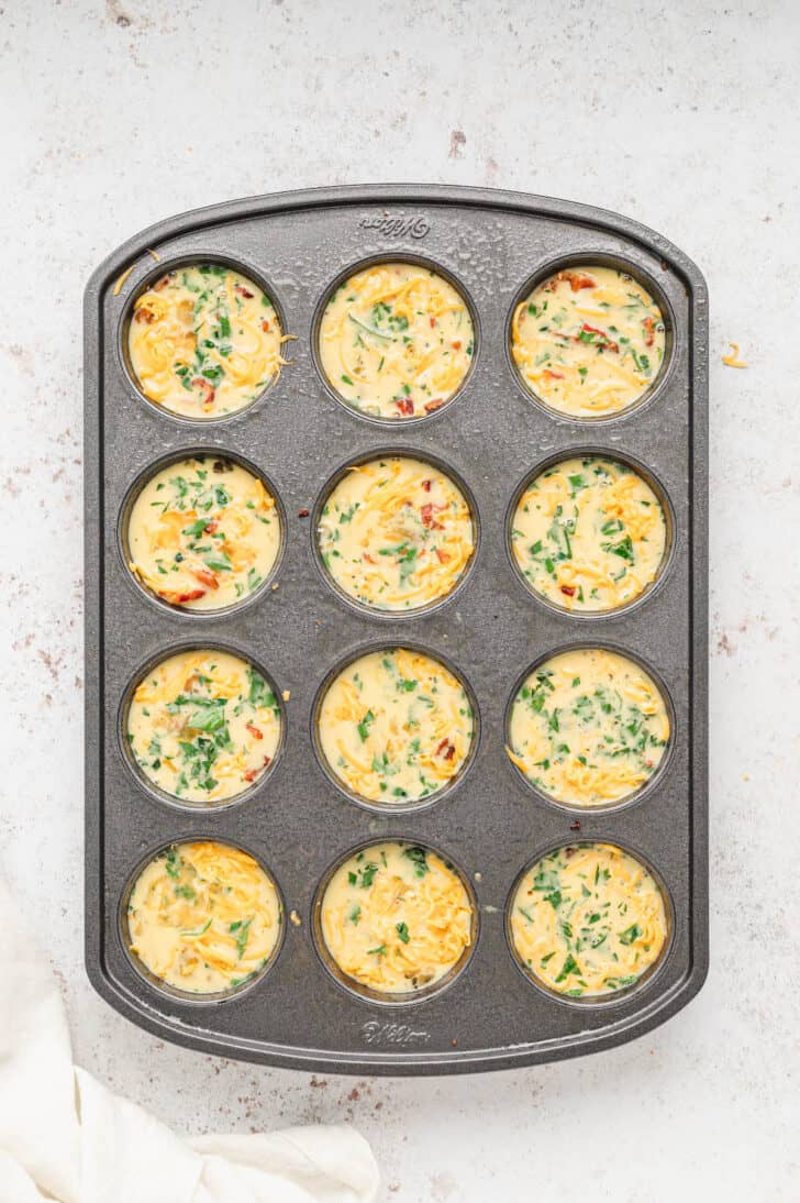A 12-cup muffin pan filled with egg muffin cups made with bacon, cheese and veggies, unbaked.