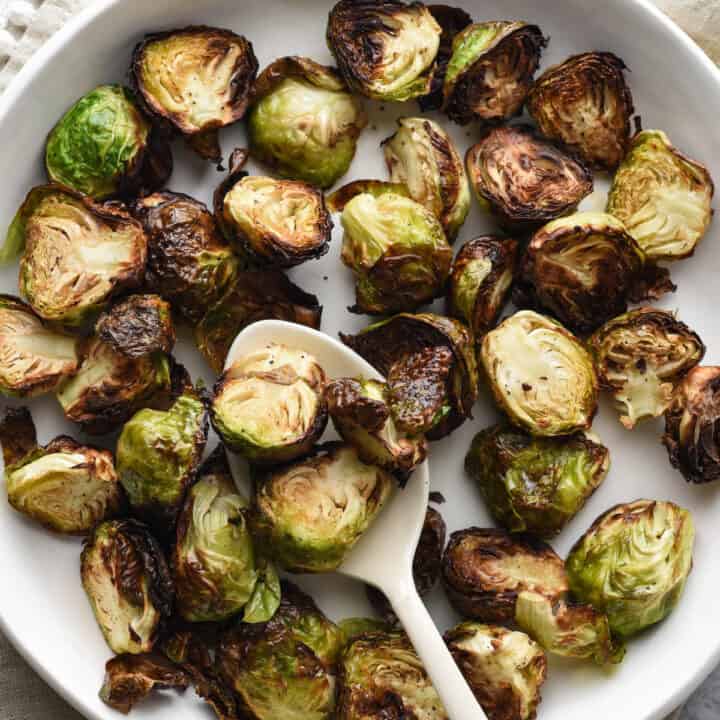 Air fried brussel sprouts in shallow white bowl with white serving spoon, on top of beige linen with pom pom edging.