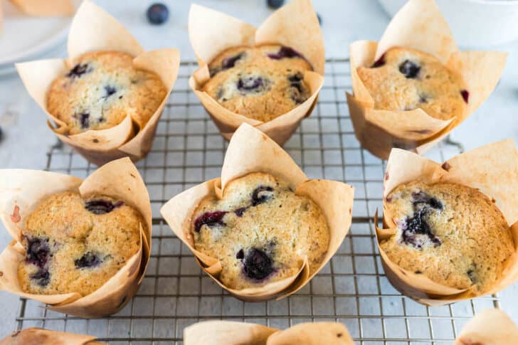 Six blueberry muffins in brown tulip paper liners on a cooling rack.