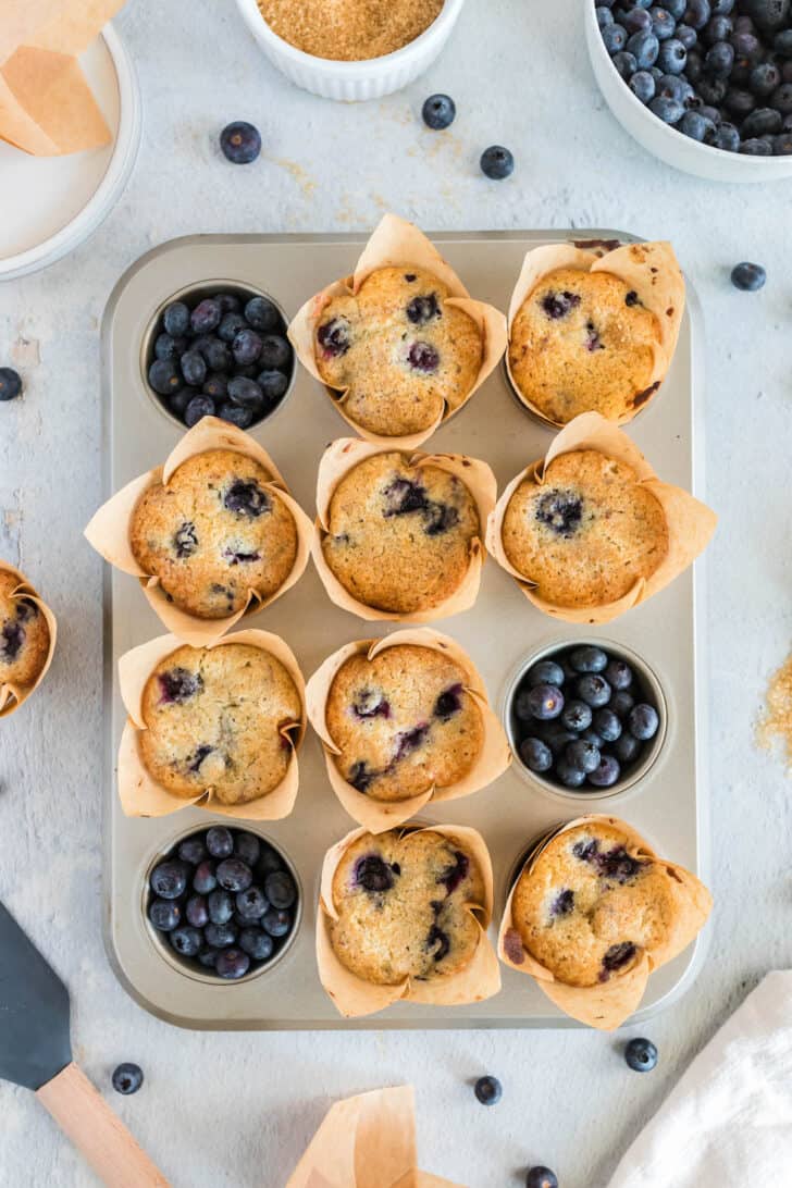Overhead photo of a muffin pan filled with 9 blueberry muffins. Fresh blueberries fill the other 3 holes.
