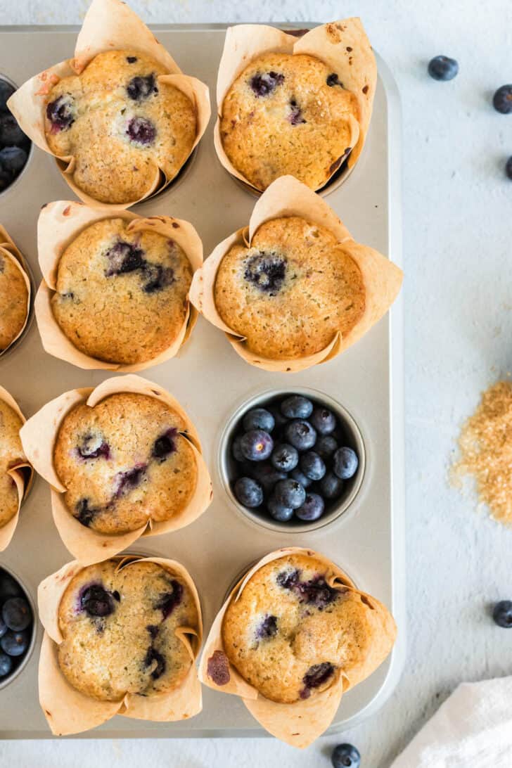 Overhead photo of 7 buttermilk blueberry muffins in a muffin pan with brown paper tulip-style liners. Fresh blueberries fill the 8th hole in the pan.