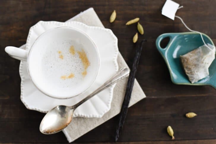 A small white teacup filled with cardamom tea latte on a matching saucer, with a spoon alongside. A vanilla bean, cardamom pods and a teabag garnish the scene.