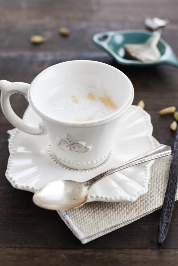 A small white teacup filled with frothy cardamom tea on a matching saucer, with a spoon alongside.