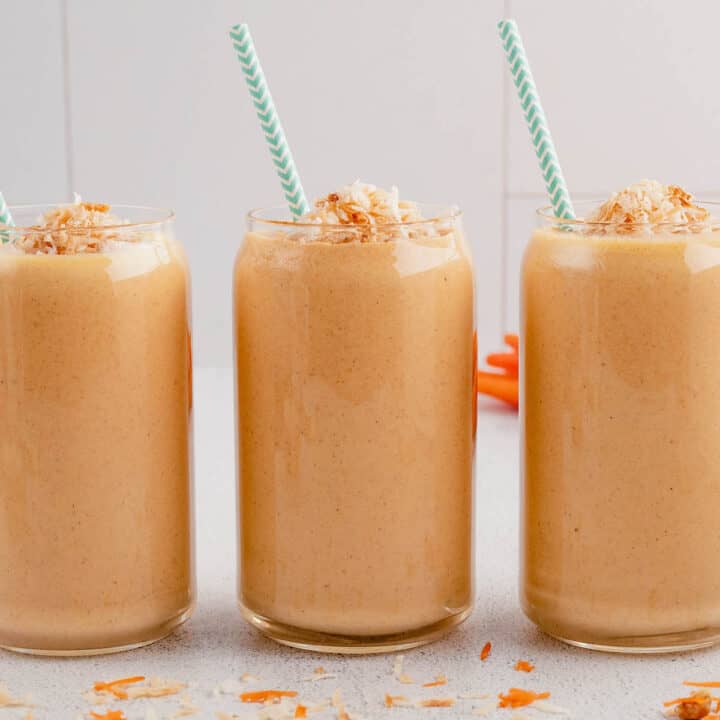 Three carrot cake smoothies lined up in a row, topped with coconut and garnished with light blue decorative paper straws.
