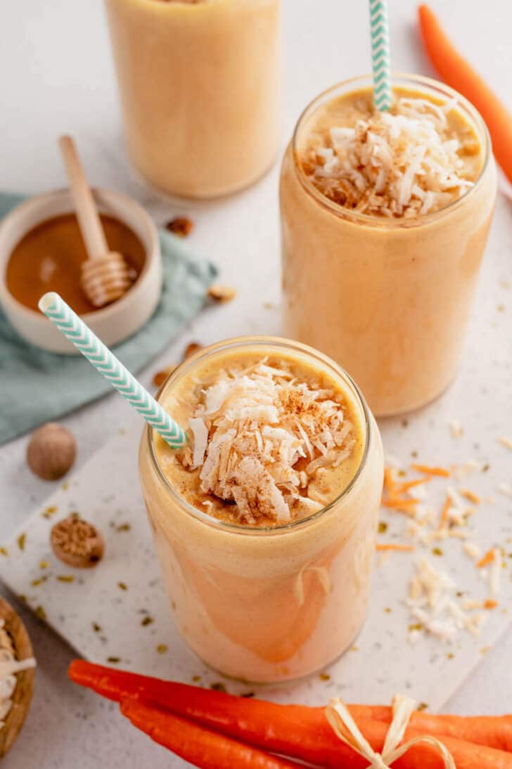 Two carrot cake smoothies topped with shredded coconut in glasses, with light blue decorative paper straws in them.