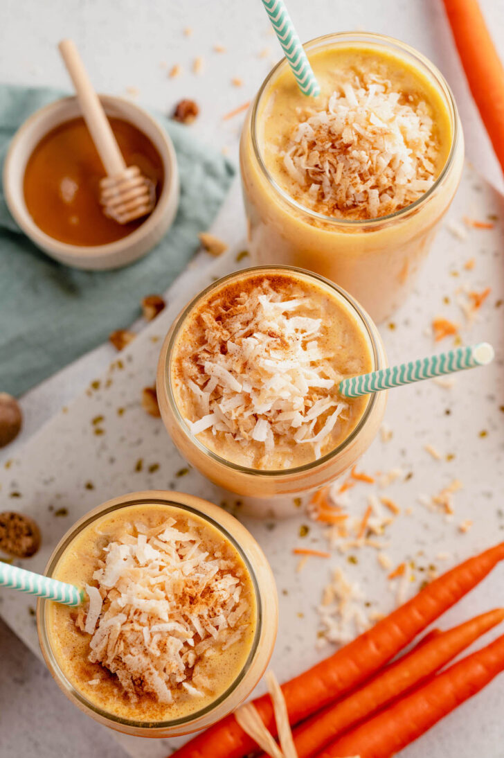 Three carrot cake smoothies topped with shredded coconut in glasses, with light blue decorative paper straws in them.