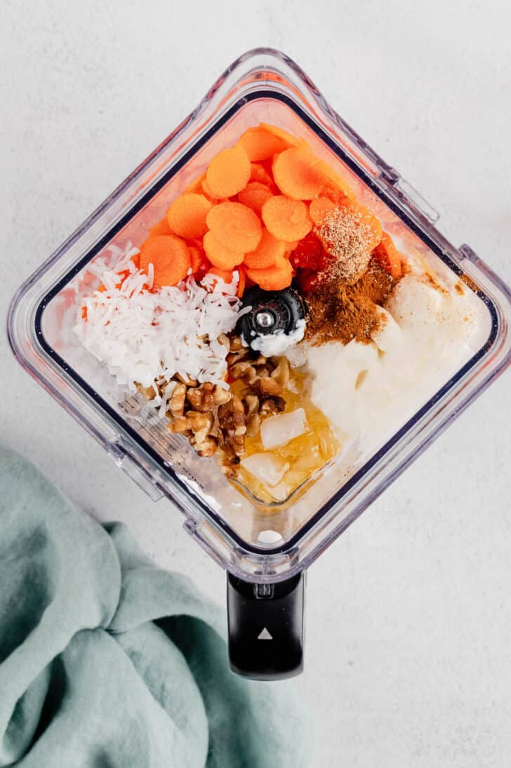 Chopped carrots, shredded coconut, walnuts, spices, yogurt, honey and ice cubes in a blender pitcher.