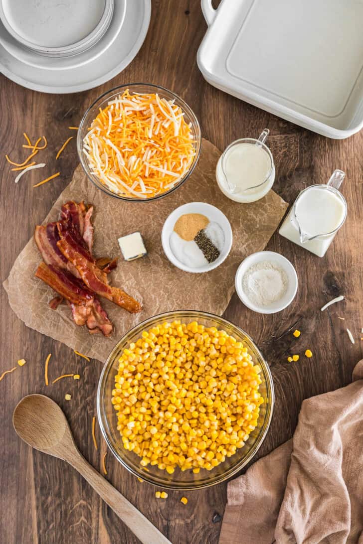 Ingredients laid out on a wooden table, including shredded Cheddar, bacon, spices, butter, flour and corn.