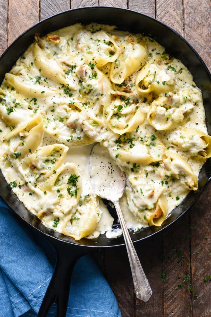 A cast iron skillet filled with chicken alfredo stuffed shells, with a spoon scooping some sauce out.
