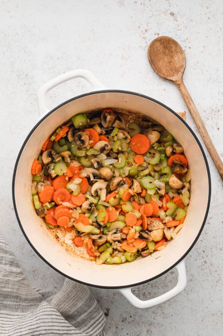 A white Dutch oven filled with sauteed carrots, celery, onions and mushrooms.