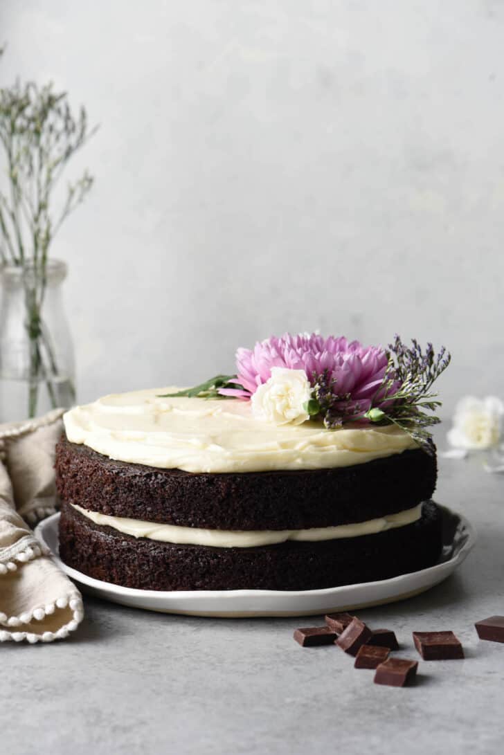 A two layer chocolate cake with cream cheese frosting on a white plate, decorated with pink and white flowers and greenery.