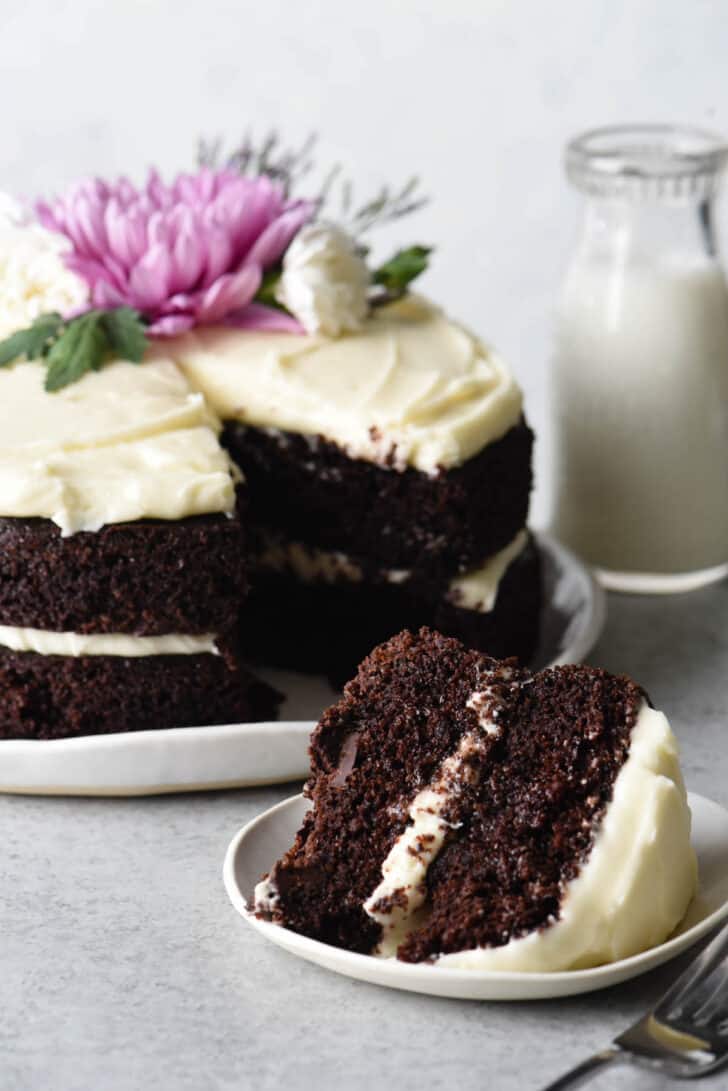 A slice of double layer chocolate cake with cream cheese frosting on a small white plate with the rest of the cake in the background.