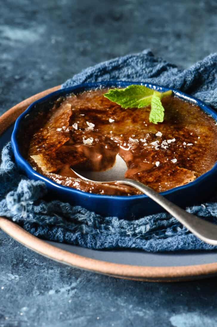 A shallow dark blue ramekin filled with chocolate creme brulee, garnished with a mint sprig and sea salt flakes, with a spoon digging into it.