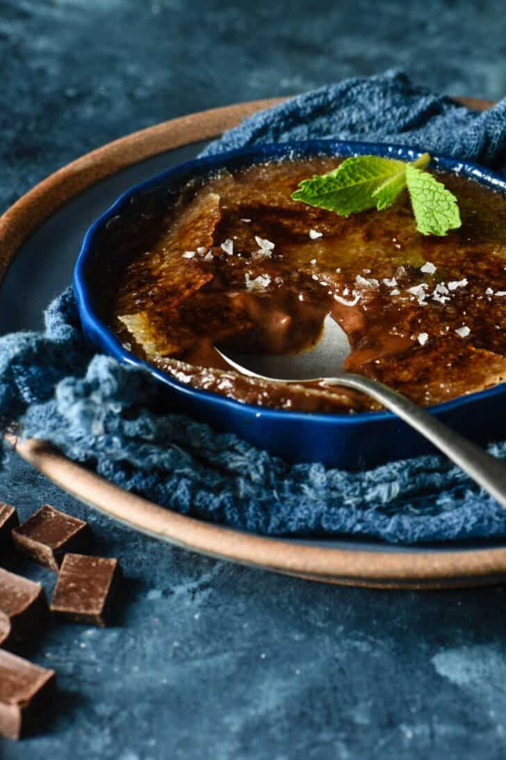 A shallow dark blue ramekin filled with dark chocolate creme brulee, garnished with a mint sprig and sea salt flakes, with a spoon digging into it.