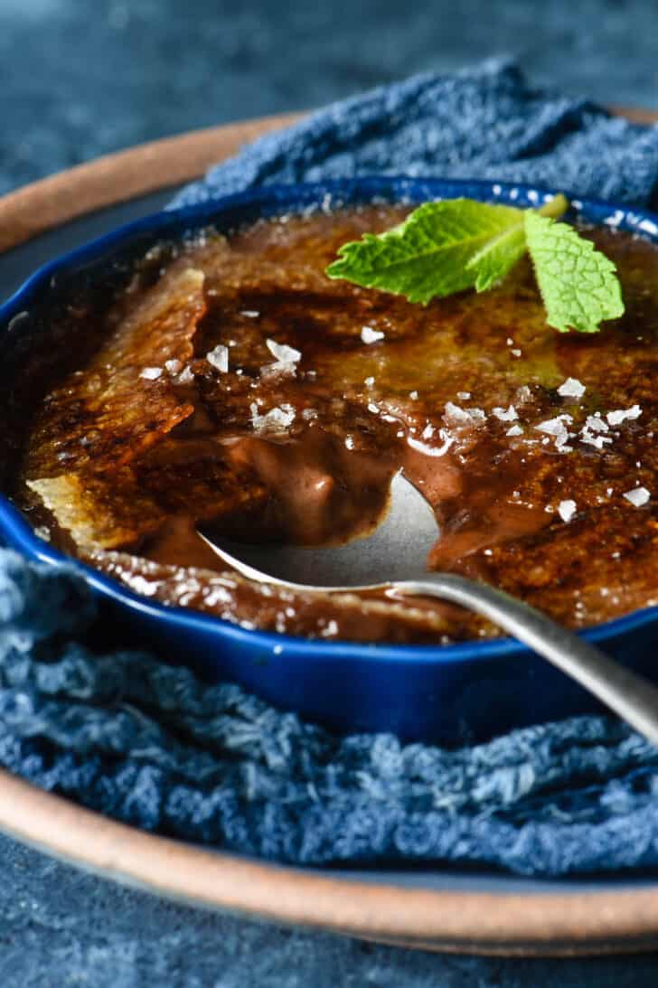 A shallow dark blue ramekin filled with dark chocolate creme brulee, garnished with a mint sprig and sea salt flakes, with a spoon digging into it.
