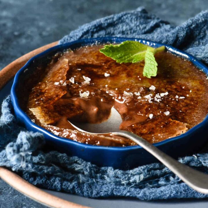 A shallow dark blue ramekin filled with chocolate creme brulee, garnished with a mint sprig and sea salt flakes, with a spoon digging into it.
